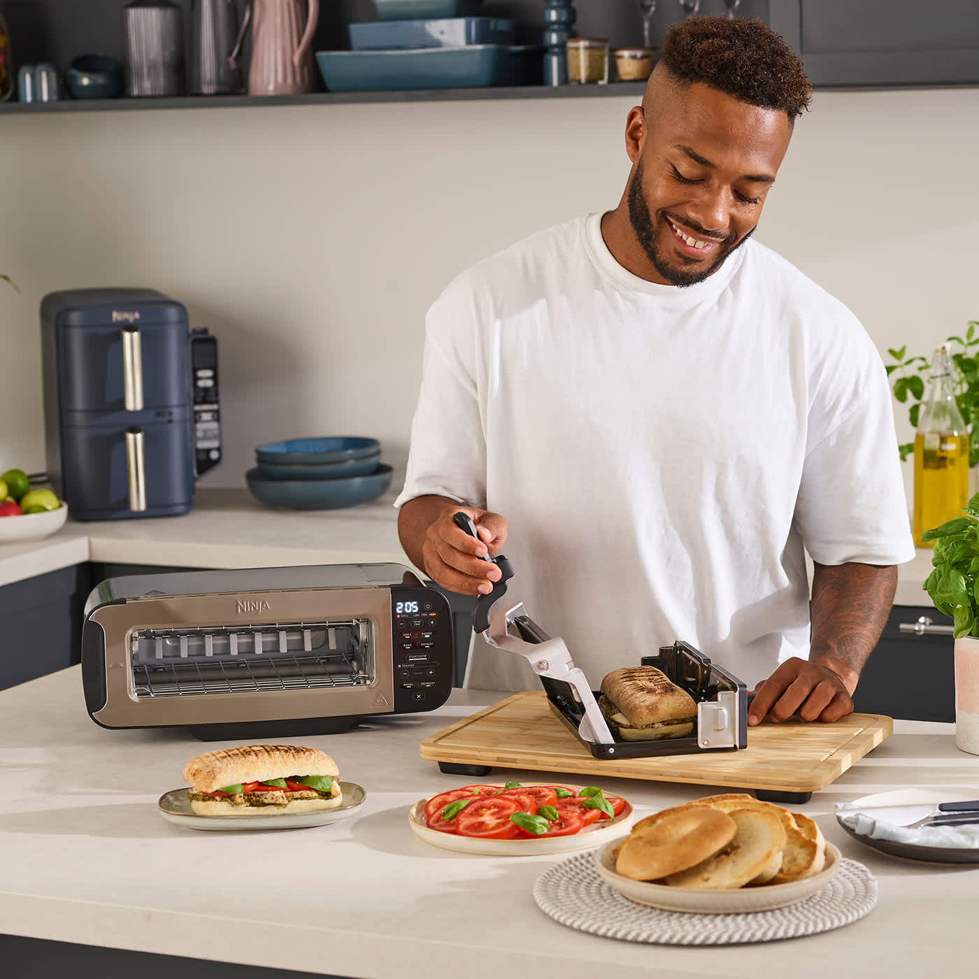 Ein Mann bereitet Paninis im Ninja Foodi 3-in-1 Gerät zu, das für die Grill- und Backfunktion horizontal umgeklappt ist. Ein Mann bereitet Paninis im Ninja Foodi 3-in-1 Gerät zu, das für die Grill- und Backfunktion horizontal umgeklappt ist.